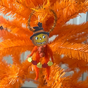 May include: A Halloween-themed ornament featuring a smiling pumpkin head wearing a black hat, attached to an orange fuzzy body. The ornament is decorated with yellow and red leaf-shaped accents and hangs from a gold tinsel ring. The background is an orange tree.