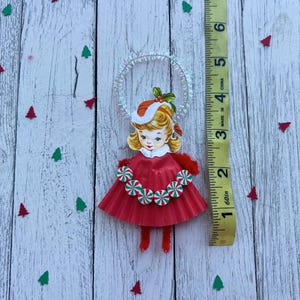 May include: A vintage Christmas ornament featuring a girl in a red dress with a candy cane trim. The ornament has a halo and is approximately 3 inches tall. The background is a white wooden surface with red and green tree-shaped confetti.