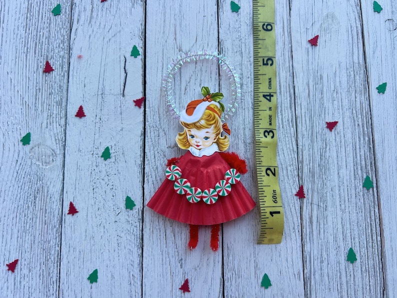 May include: A vintage Christmas ornament featuring a girl in a red dress with a candy cane trim. The ornament has a halo and is approximately 3 inches tall. The background is a white wooden surface with red and green tree-shaped confetti.