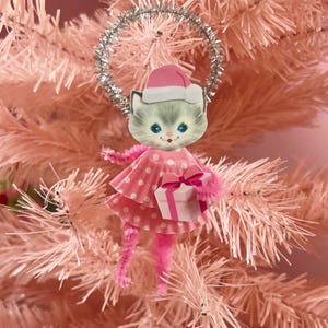 May include: A holiday ornament featuring a cat face with a pink Santa hat, a pink polka-dot cupcake liner skirt, and pink pipe cleaner limbs. The cat holds a small white gift box with a pink ribbon, and a silver tinsel halo sits above the head.