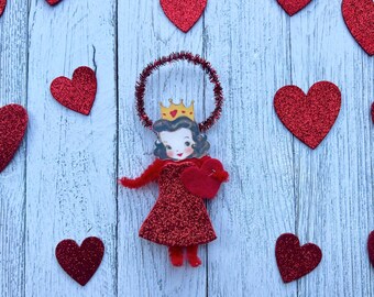 May include: A vintage-style Valentine's Day ornament featuring a woman in a red glitter dress and a gold crown, holding a red heart. The ornament is surrounded by red glitter heart decorations on a light blue wooden background.