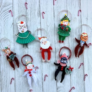 May include: A collection of vintage-style Christmas ornaments. The ornaments include a cat, Santa Claus, a girl with a Christmas tree hat, a gingerbread man, a reindeer, a snowman, and a black dog. Each ornament has a wire loop for hanging and is decorated with candy canes.