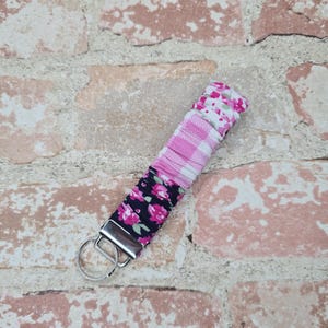 May include: A fabric keychain with a metal ring. The keychain is made of several patterned fabrics: pink gingham, white with pink flowers, and navy blue with pink roses. The keychain is designed to hold keys.