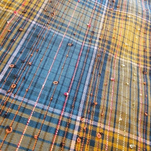 May include: Close-up of a blue and yellow plaid fabric with a woven texture. The fabric has a subtle sheen and is decorated with small, evenly spaced brown dots.