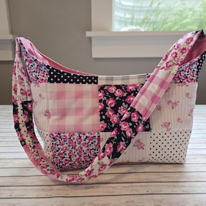 May include: A patchwork shoulder bag with a variety of floral, gingham, and polka dot patterns in pink, white, and navy blue. The bag has a long strap and is made of quilted fabric. The bag is sitting on a light wood surface.