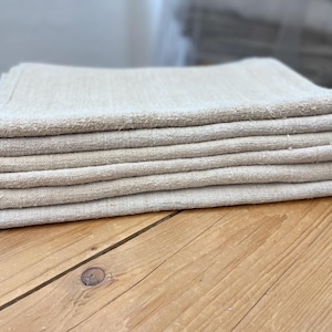 May include: A stack of folded, beige linen napkins. The napkins are neatly arranged on a wooden surface, showcasing their textured weave. The neutral color and simple design suggest a rustic or farmhouse aesthetic, suitable for kitchen or dining decor.