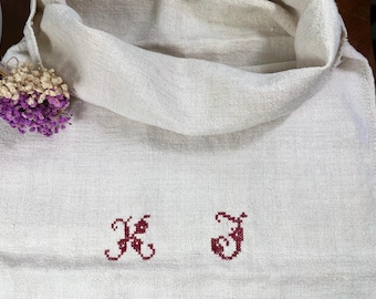 May include: Cream-colored linen towel with embroidered initials "K J" in burgundy thread. A small bundle of dried purple and white flowers rests on the upper left corner. The towel is folded and draped.