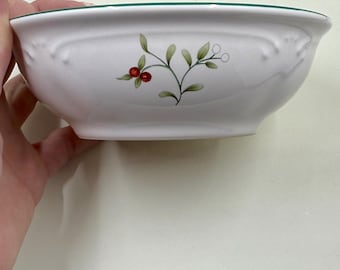 May include: White ceramic bowl with a green rim and a floral design of red berries and green leaves. The bowl has a curved shape. The bowl is likely for serving food. The bowl is held by a hand.