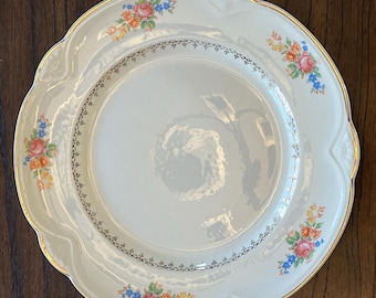 May include: A white ceramic dinner plate with a gold rim and floral pattern. The plate has a scalloped edge and a delicate floral design in shades of pink, blue, and yellow.