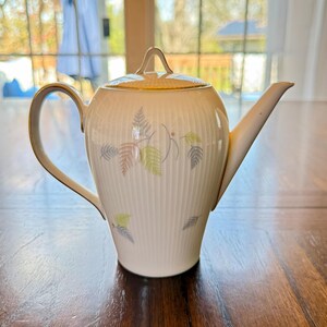 May include: Cream-colored ceramic teapot with a lid and a long spout. The teapot features a gold handle and trim, and is decorated with a pattern of stylized fern leaves in shades of green, brown, and gray. The teapot is sitting on a wooden surface.