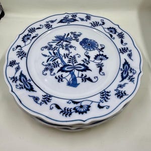 May include: A stack of white ceramic plates with a blue floral design. The plates have a scalloped edge and a detailed blue pattern featuring flowers and foliage. The plates are stacked on top of each other.