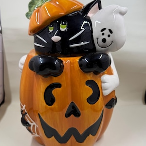 Op de afbeelding: Een keramieken koekjespot in de vorm van een grote oranje pompoen met een uitgesneden jack-o'-lantern gezicht. Een zwarte kat met een klein pompoenhoedje en een wit spookje zitten bovenop. Een kleine zwarte spin in een wit web is zichtbaar aan de zijkant.