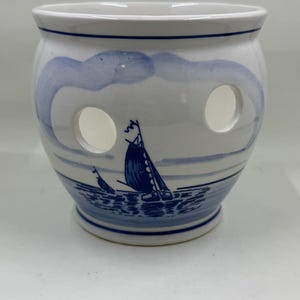 May include: A white ceramic planter with a blue nautical design. The planter features a sailboat scene and two circular openings. The rim and base are accented with blue lines, creating a classic, coastal aesthetic.