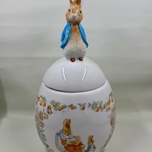 Pode incluir: Um pote de cerâmica branco com tampa, encimado por uma figura de Peter Rabbit. O pote é adornado com ilustrações de coelhos e padrões florais. A figura do coelho usa um casaco azul.