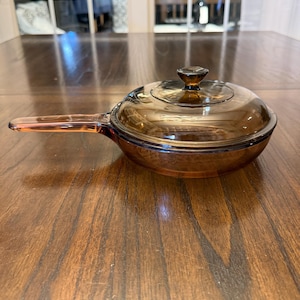 Puede incluir: Una cacerola de vidrio ámbar vintage con tapa a juego. La cacerola tiene un mango largo y una tapa de vidrio transparente con un pequeño pomo. La batería de cocina está sobre una superficie de madera.