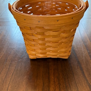May include: A round, light brown woven wood basket featuring a wide, reinforced rim with visible metal rivets. The basket has a tapered shape with a flat base and two small, integrated wooden handles on opposite sides of the upper rim.