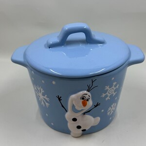 May include: A light blue ceramic pot with a matching lid. The pot features a raised image of Olaf from Frozen, along with snowflake designs. The pot has two side handles and a handle on the lid.