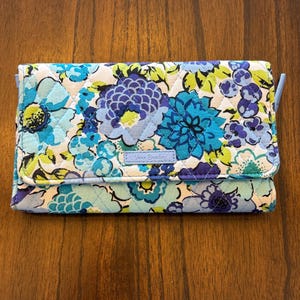 May include: A quilted wallet with a floral design in shades of blue, teal, and white. The wallet has a flap closure and a small blue strap. The brand name "Vera Bradley" is visible on the front.