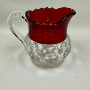 Puede incluir: Una jarra de crema de vidrio transparente con la parte superior y el asa de color rojo rubí. El borde superior tiene un borde festoneado con un ribete verde oscuro. La parte inferior de la jarra de crema tiene un diseño claro y texturizado. El asa es de vidrio transparente.
