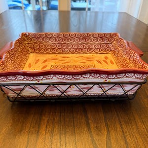 May include: Rectangular ceramic baking dish with a red and cream floral pattern, set in a black metal wire basket. The dish has red handles and a decorative rim. The interior is a light yellow color.