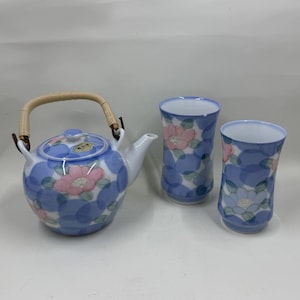 Puede incluir: Juego de té de cerámica azul y blanco con una tetera y dos tazas a juego. La tetera tiene un asa tejida y un diseño floral con flores rosas y blancas. Las tazas también presentan el mismo patrón floral.