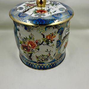 Puede incluir: Un bote cilíndrico de metal con tapa extraíble que presenta un pomo esférico de color dorado. El recipiente está decorado con paneles blancos, bordes azules ornamentados e ilustraciones florales coloridas de flores naranjas, rosas y amarillas.