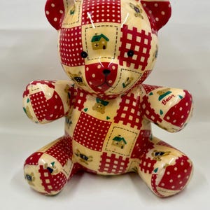 May include: A decorative teddy bear figurine with a patchwork design. The bear features red and cream-colored patches with polka dots, crosshatch patterns, and small house and bear motifs. The word "Bear" is written on one of the bear's paws.