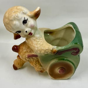 May include: A vintage ceramic planter shaped like a lamb pulling a green cart. The lamb has a cream-colored body, brown ears, and a pink nose. The cart has a green exterior with brown wheels and accents. A charming decorative piece.