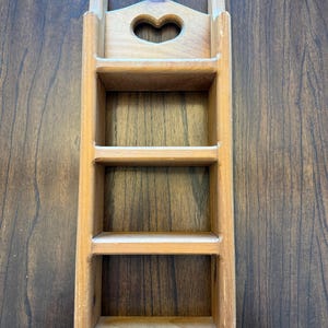 May include: A light-colored wooden wall shelf with three compartments. The top features a heart-shaped cutout. The shelf is rectangular with two vertical supports and a handle-like top. The wood grain is visible.