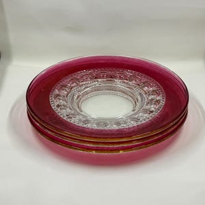 May include: A stack of four glass plates with a ruby red rim and a clear, ornate center. The plates have a gold trim and are stacked on top of each other. The plates are on a white surface.