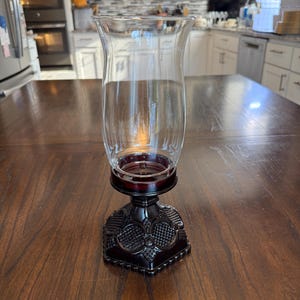 May include: A clear glass hurricane candle holder featuring a removable top cylinder and a dark red, hexagonal pressed glass base with a textured diamond and fan pattern. The base supports the glass insert with a circular rim.