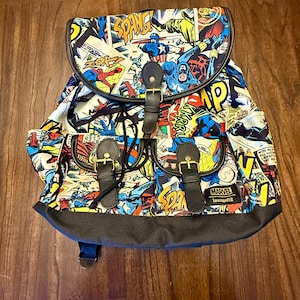 May include: A Marvel comic book-themed backpack with a flap closure and two front pockets. The backpack features a colorful collage of comic book panels with characters like Spider-Man and Captain America. It has black straps and buckles, and the Marvel Loungefly logo.