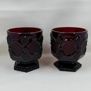 May include: A pair of deep red glass tumblers featuring a raised geometric pattern with diamond-shaped textures and circular motifs. Each tumbler has a wide, open mouth and sits on a sturdy, hexagonal-shaped base, showcasing a rich, dark crimson hue.