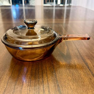 Puede incluir: Cacerola de vidrio color ámbar con tapa y asa. La batería de cocina tiene forma redondeada y está hecha de vidrio transparente, lo que permite ver el contenido. El asa es una extensión del cuerpo de la cacerola.