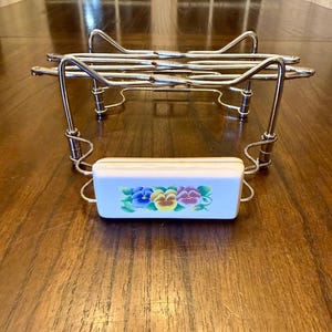 May include: A silver-toned metal serving stand with a white rectangular dish. The dish is decorated with colorful pansies. The stand has a wire frame design and is designed to hold the dish.