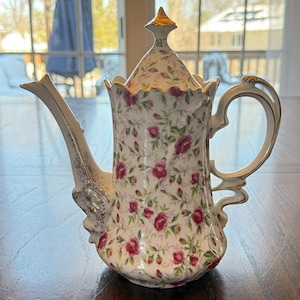 May include: A white porcelain teapot with a floral pattern of pink roses and green leaves. The teapot has a gold-colored handle, spout, and lid accents. The lid is topped with a gold finial. The teapot is on a wooden table.