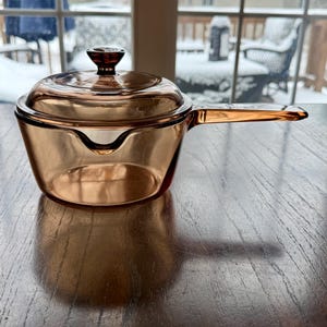 Puede incluir: Una cacerola de vidrio ámbar vintage con tapa y asa a juego. La cacerola tiene un cuerpo redondeado y un asa larga. La tapa tiene un pequeño pomo. El vidrio es transparente. La cacerola está sobre una superficie de madera.