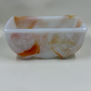 Op de afbeelding: Een rechthoekige, witte glazen schaal met oranje en rode marmering. De schaal heeft een bloemenmotief aan de zijkanten en is waarschijnlijk vintage. De schaal is ongeveer 15 cm lang, 10 cm breed en 8 cm hoog.