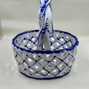 May include: White ceramic basket with a blue lattice design and a twisted handle. The rim and handle are decorated with blue floral patterns. The basket has a round shape and is likely used for decorative purposes.