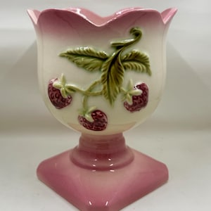 May include: A ceramic pedestal vase with a scalloped rim, featuring a cream-colored body with a pink gradient at the top and base. The front is decorated with a raised, three-dimensional relief of green leaves and three dark red strawberries.