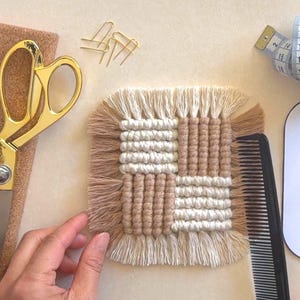 Puede incluir: Un posavasos de macram&eacute; hecho a mano en colores beige y crema. El posavasos cuadrado presenta un dise&ntilde;o tejido con un borde con flecos. Tambi&eacute;n se ven tijeras doradas, clips, un peine y una cinta m&eacute;trica.