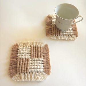 Puede incluir: Dos posavasos cuadrados de macram&eacute; en beige y marr&oacute;n claro con un borde con flecos. Un posavasos tiene una taza de t&eacute; blanca encima. Los posavasos tienen un dise&ntilde;o tejido con un patr&oacute;n de cuadr&iacute;cula. Los posavasos est&aacute;n hechos a mano.