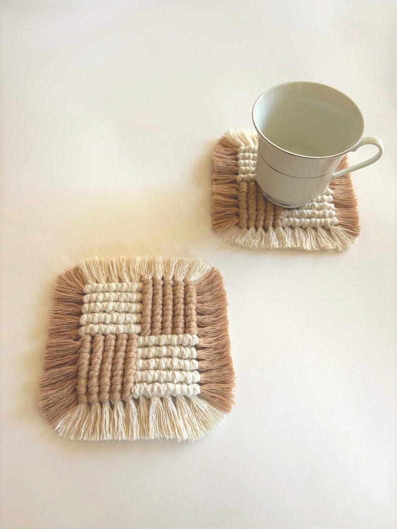 Puede incluir: Dos posavasos cuadrados de macram&eacute; en beige y marr&oacute;n claro con un borde con flecos. Un posavasos tiene una taza de t&eacute; blanca encima. Los posavasos tienen un dise&ntilde;o tejido con un patr&oacute;n de cuadr&iacute;cula. Los posavasos est&aacute;n hechos a mano.