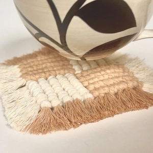 Puede incluir: Un posavasos cuadrado de macram&eacute; en colores beige, crema y marr&oacute;n claro. El posavasos tiene un dise&ntilde;o tejido con un borde con flecos. Una taza de cer&aacute;mica estampada se coloca encima.