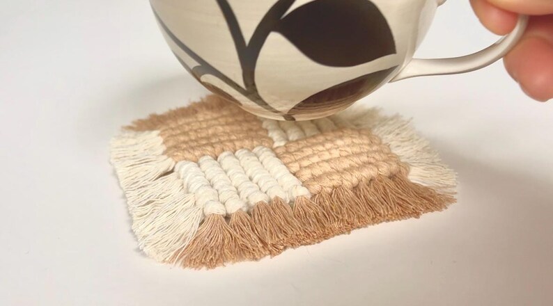 Puede incluir: Un posavasos cuadrado de macram&eacute; en colores beige, crema y marr&oacute;n claro. El posavasos tiene un dise&ntilde;o tejido con un borde con flecos. Una taza de cer&aacute;mica estampada se coloca encima.