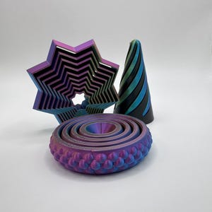 May include: A collection of iridescent 3D-printed objects. The objects include a star-shaped design, a spiral cone, and a circular, layered design with a textured edge. The colors shift from purple to blue and green.