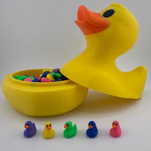 May include: A large yellow duck-shaped container with an open lid, revealing colorful candies inside. Five small, colorful duck figurines are lined up in front of the container. The duck's beak is orange, and it has a black eye.