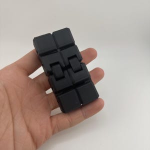 May include: A black fidget toy, held in a hand, composed of connected, square blocks. The toy is made of a matte material and has a simple, geometric design. The blocks are connected by hinges, allowing for movement and manipulation.