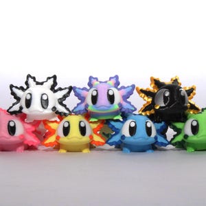 May include: A collection of colorful, cartoonish axolotl figurines. Each figure has a unique color scheme, including pink, white, yellow, blue, green, and black, with contrasting gill-like structures. The figurines have large, expressive eyes.