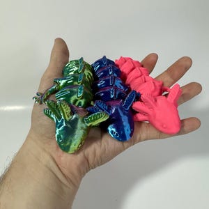 May include: A hand holding four colorful, articulated axolotl figurines. The axolotls are in shades of green, blue, and pink, with a metallic sheen. The figurines are detailed and appear to be 3D-printed.